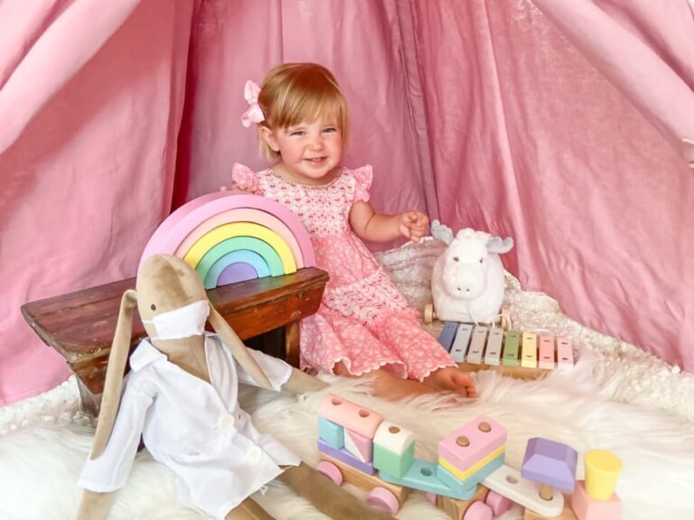 Little toddler girl in a pink dress playing on a fluffy rug with a wooden train and a wooden rainbow