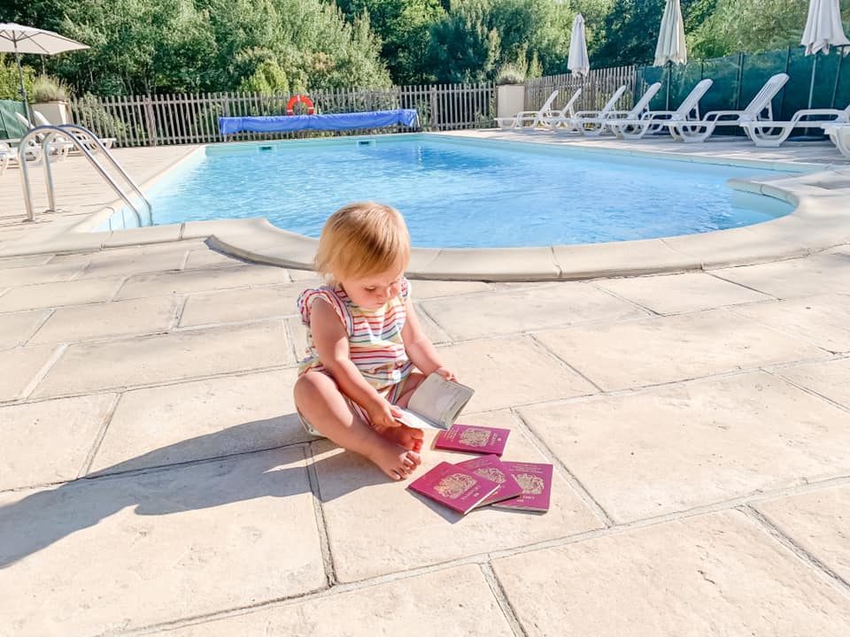 baby sat on patio alongside a swimming pool in striped top looking in a passport surrounded by four more closed British passports