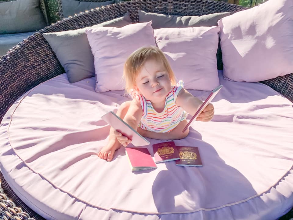 baby in striped top holding passport on a big round pink sun chair surrounded by pin and grey cushions