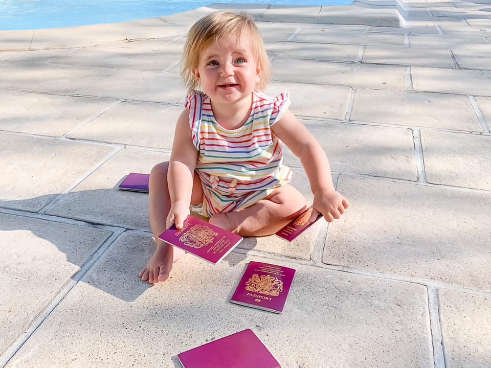 baby in striped top sat on the floor beside a pool outside holding multiple passports 