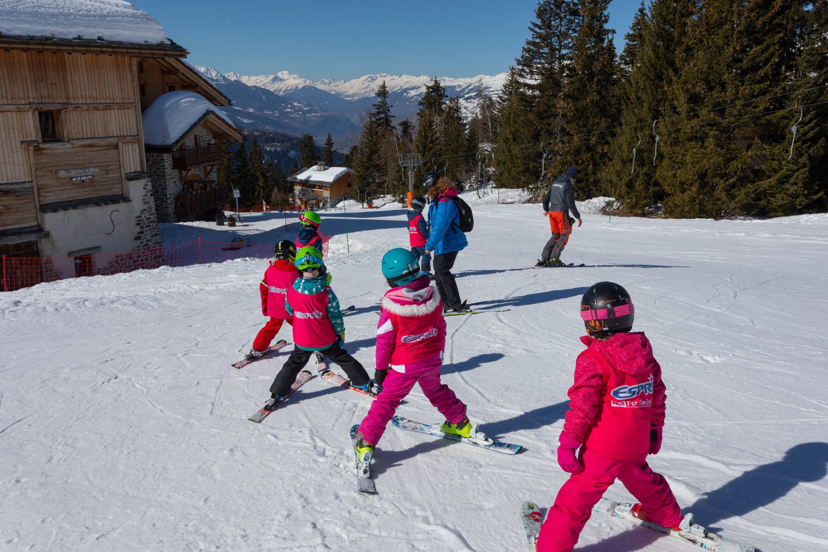 children on the snow at ski practice all wearing pink jackets and trousers with green, black and blue helmets listening to adults wearing blue jacket and backpack