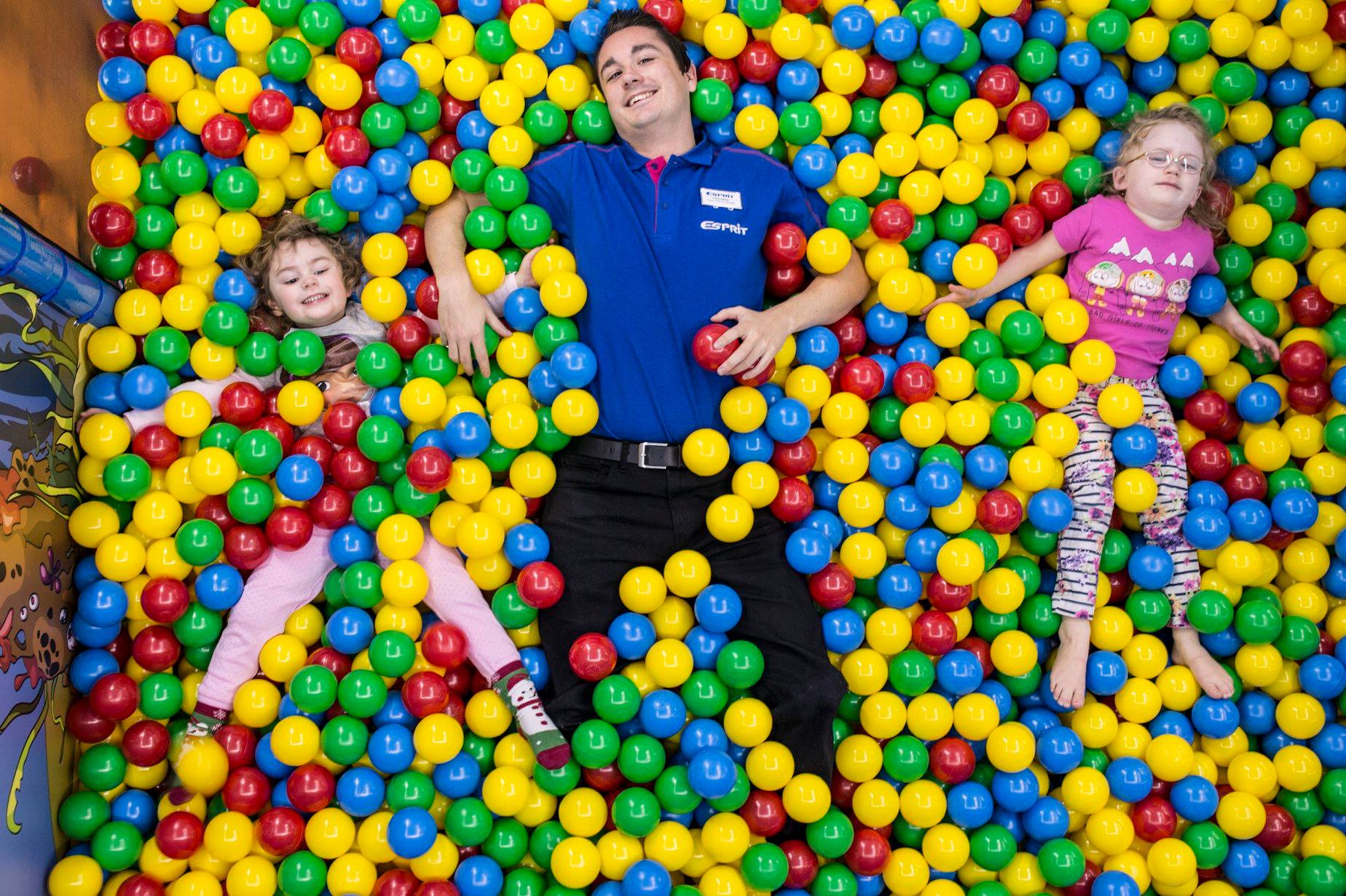 picture of esprit children's entertainer wearing a blue t-shirt and black trousers laid in a ball pool looking up to the camera along side two girls wearing pink also laid on their back in a multi coloured ball pit