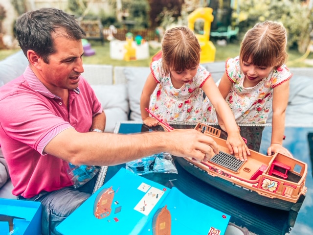 twin girls and daddy in the garden building a playmobil pirate ship. The twins are passing daddy pieces to help put it together 