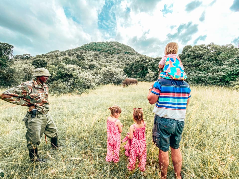 family with child on shoulders of father and two twins wearing pink jumpsuits along side safari ranger overlooking the rhino in and amongst knee high grass surrounded by trees