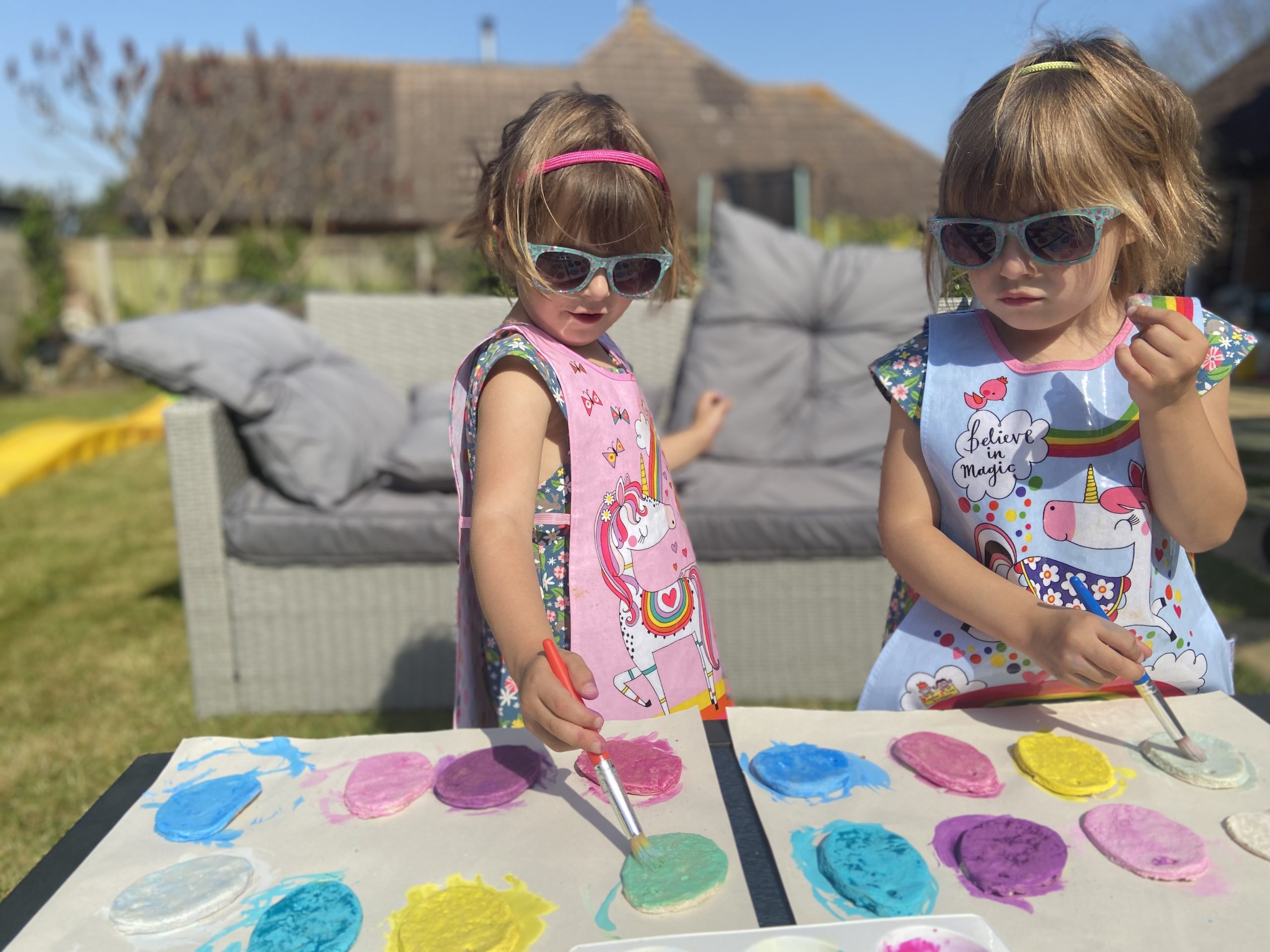 two girls outside in the garden wearing art aprons painting salt dough with a paintbrush