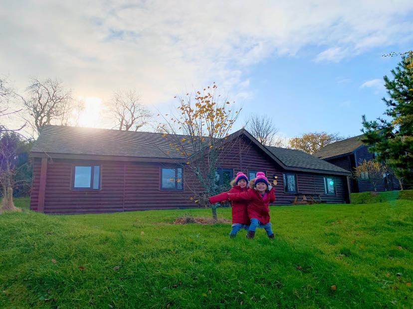 Two children in red coats and bobble hats standing out the front of a wooden lodge, on the grass at Bluestone in Wales 