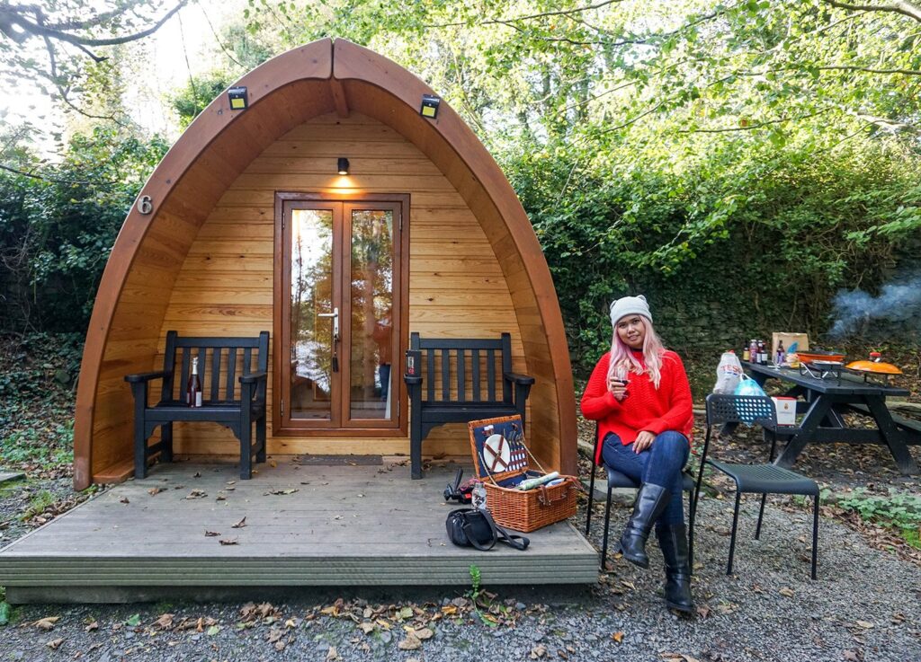 wooden wigwam with a small decking out of the front, and two wooden chairs placed either side of the door. to the right of the wigwam is a lady wearing a red coat sitting on a chair