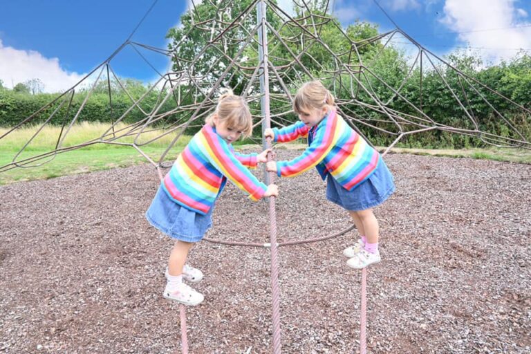 two girls balancing on a spiders web climbing frame waring blue skirts and rainbow jumpers