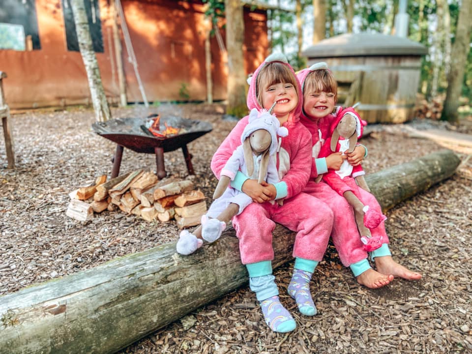 two children sitting on a log with a campfire lit behind them and the hot tub in the distance 