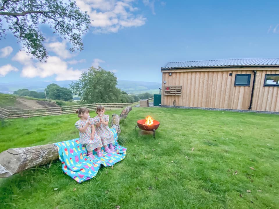 two girls sitting on a log dating marshmallows with a small fire going next to them looking out over the hills in wales