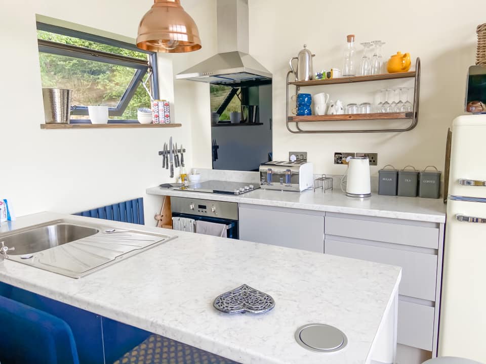 The kitchen at Penlan lodges with a white worktop, silver sink and cooker with shelves up on the wall