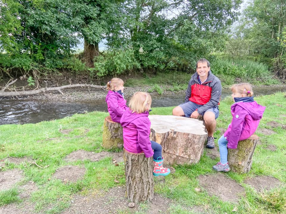 three young girls in purple coats and a daddy sat on logs besides a river