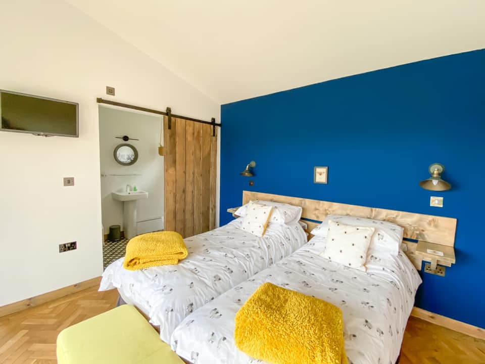 Twin bedroom at Penlan lodges with sheep beeding and mustard blankets on the bottom of the bed. to the left is an ensuite bathroom with the door open. 