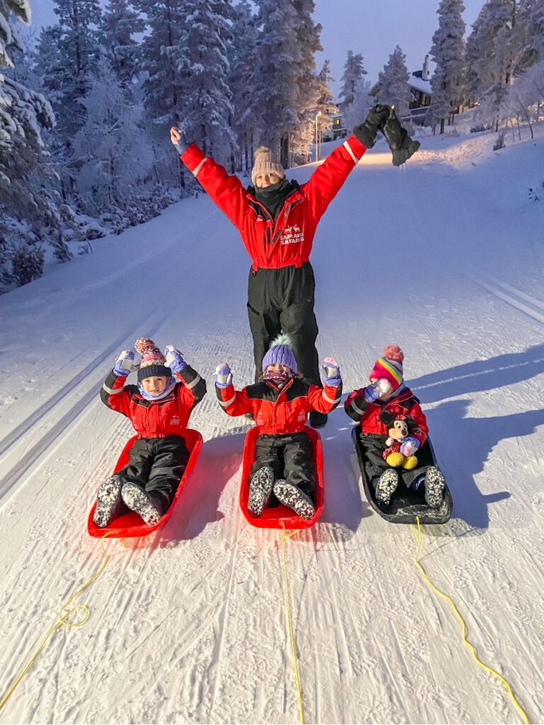 three children sledging in the snow with an adult cheering with her hands in the air - Lapland Holiday 